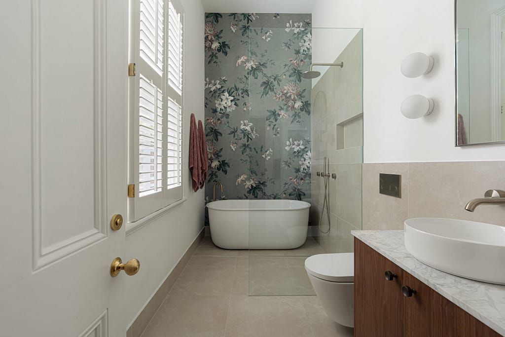 Modern bathroom featuring a freestanding bath against floral wall tiles, walk-in shower with brushed nickel fixtures, plantation shutters, and a timber vanity with marble top.